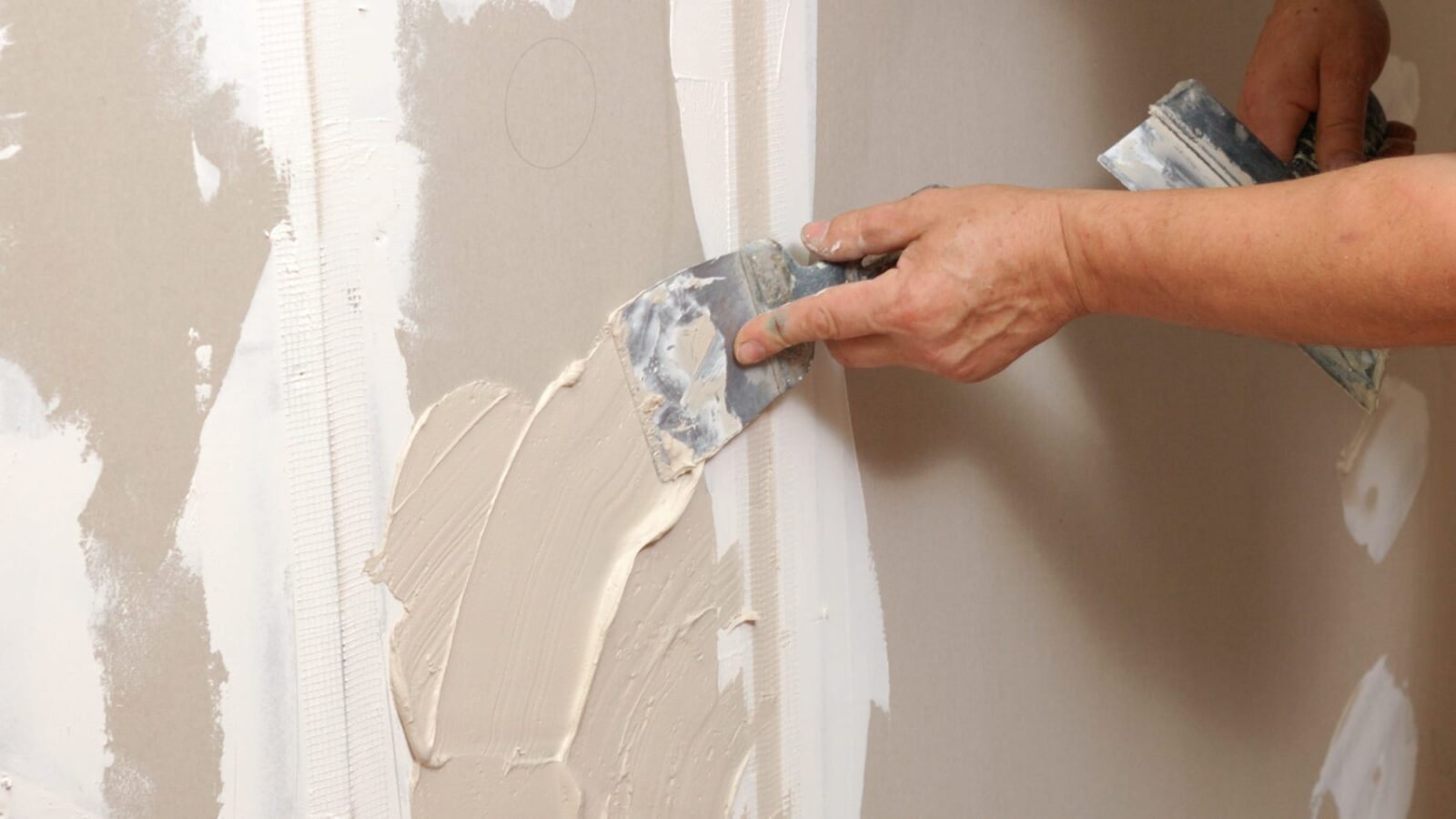 A close-up shows a contractor using a drywall knife to smoothly apply beige joint compound over the wall and mesh tape.