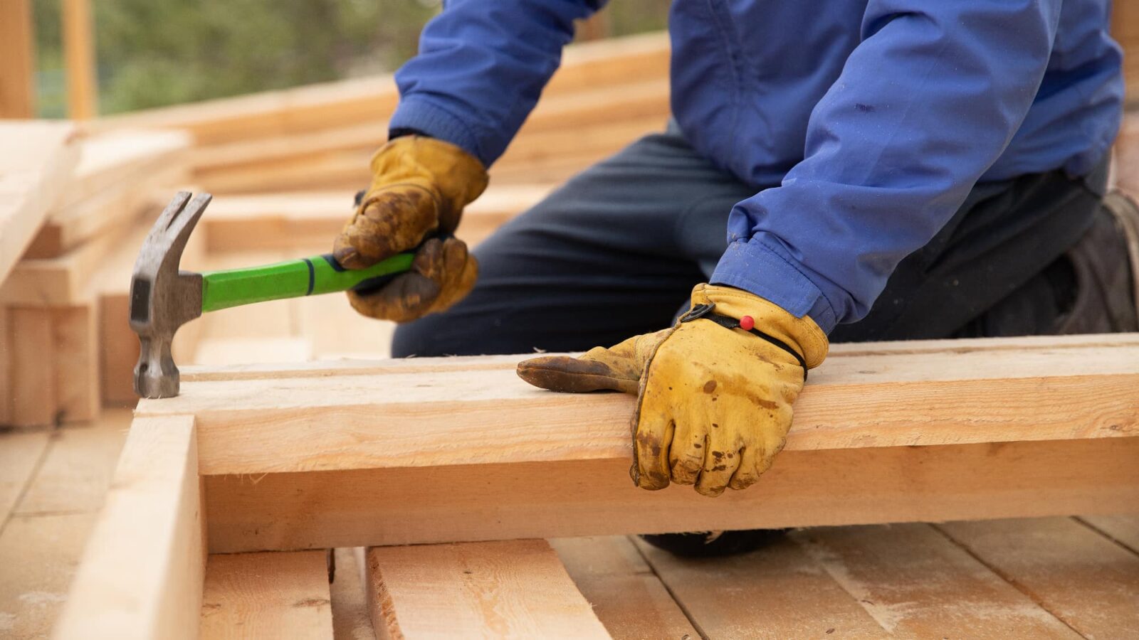 A construction worker in a blue jacket and yellow gloves is kneeling and using a hammer with a green handle to nail two pieces of light-colored wood together.