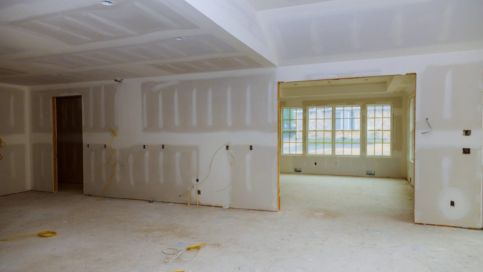 An interior space undergoing renovation with freshly installed drywall walls and ceiling, showing seam compound and exposed yellow electrical wires.