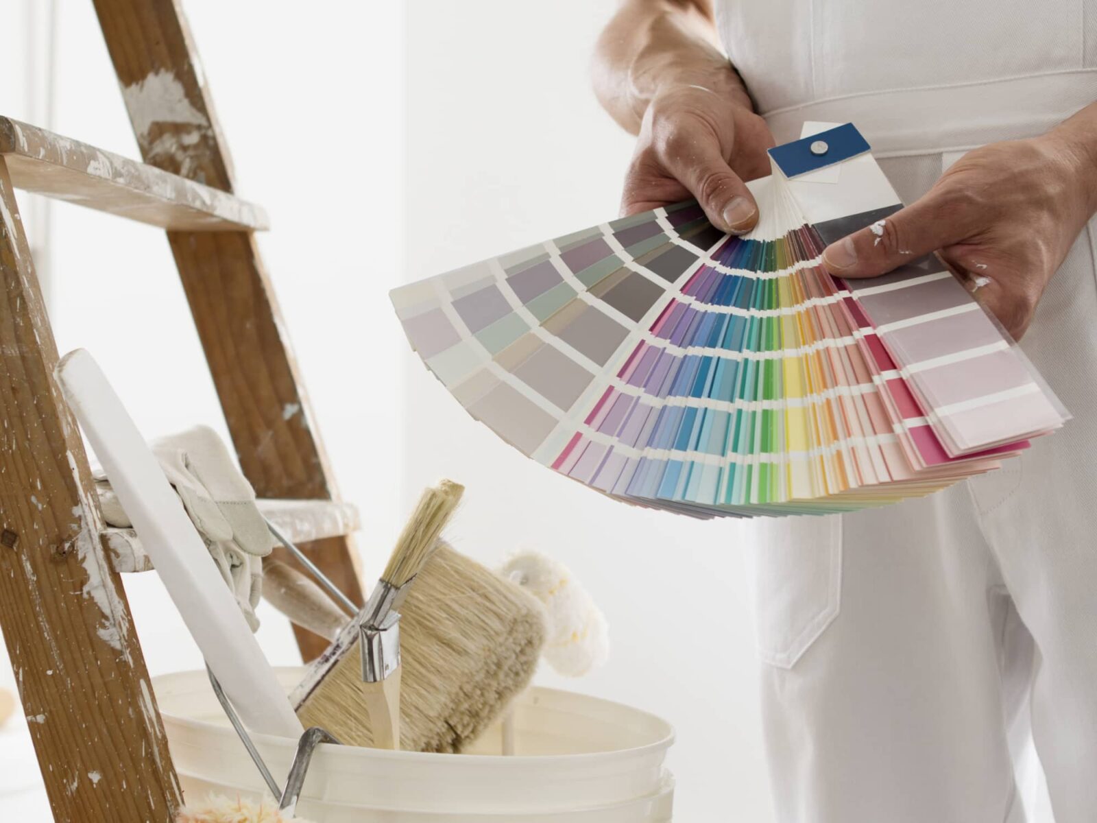A painter in white overalls holds a large fan of multi-colored paint swatches. Nearby is a ladder and a bucket with brushes.