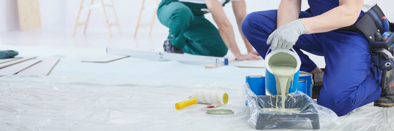 A painter in blue overalls pours light green paint from a bucket into a roller tray on a plastic-covered floor, with another worker nearby.