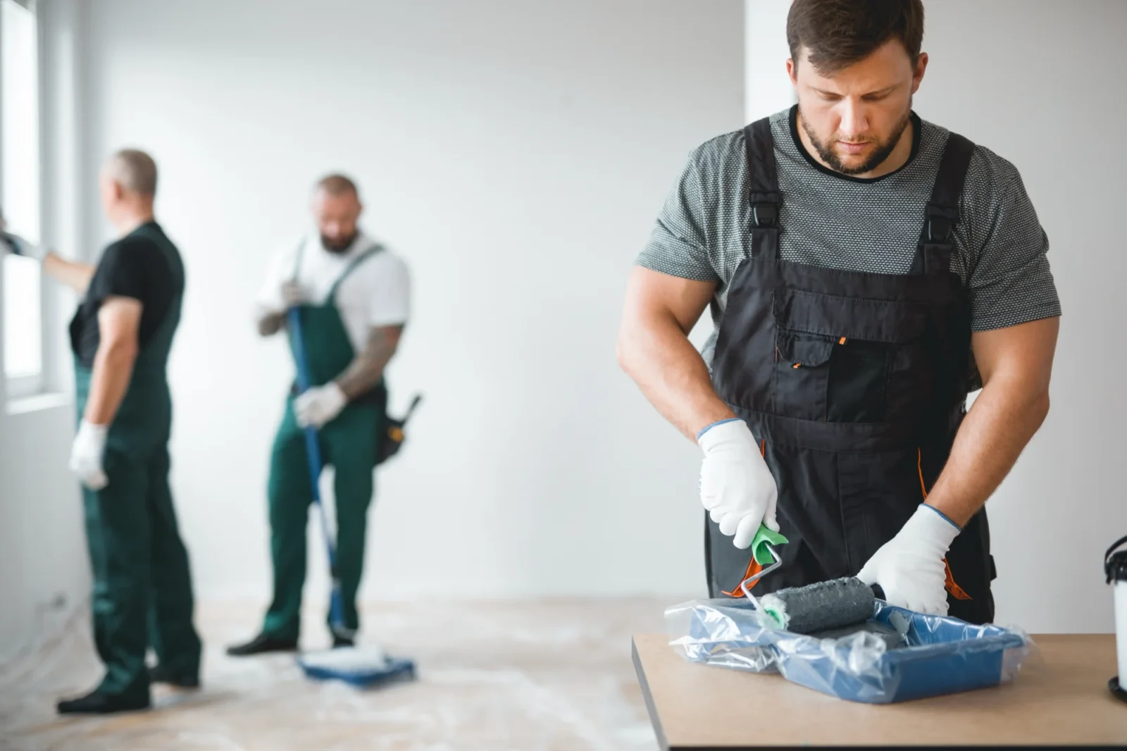 A professional painter in grey overalls prepares a roller with paint in the foreground. Two colleagues in green overalls work on the walls of the bright interior room behind him.